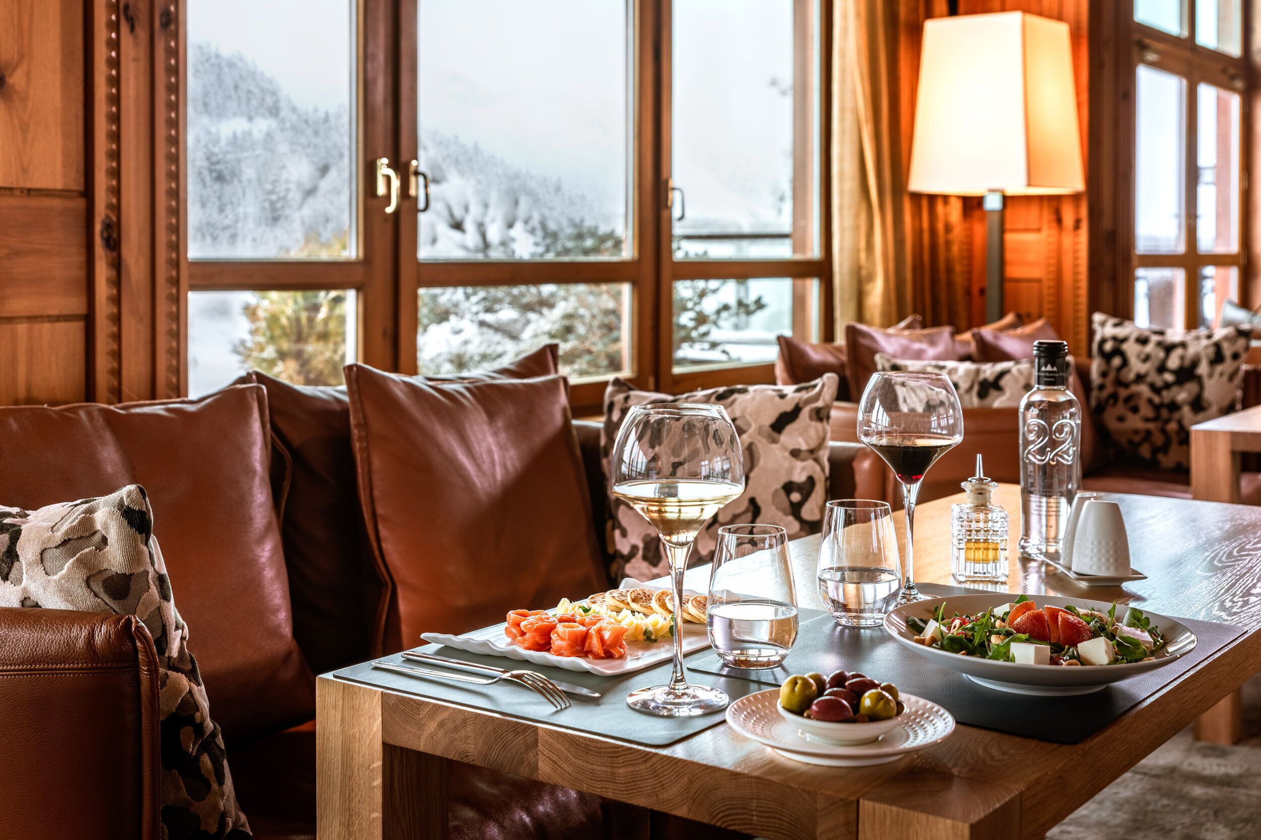 A cozy dining area with a wooden table set for a meal, a sofa with leather cushions, and a view of snowy mountains through large windows.