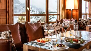 A cozy dining area with a wooden table set for a meal, a sofa with leather cushions, and a view of snowy mountains through large windows.