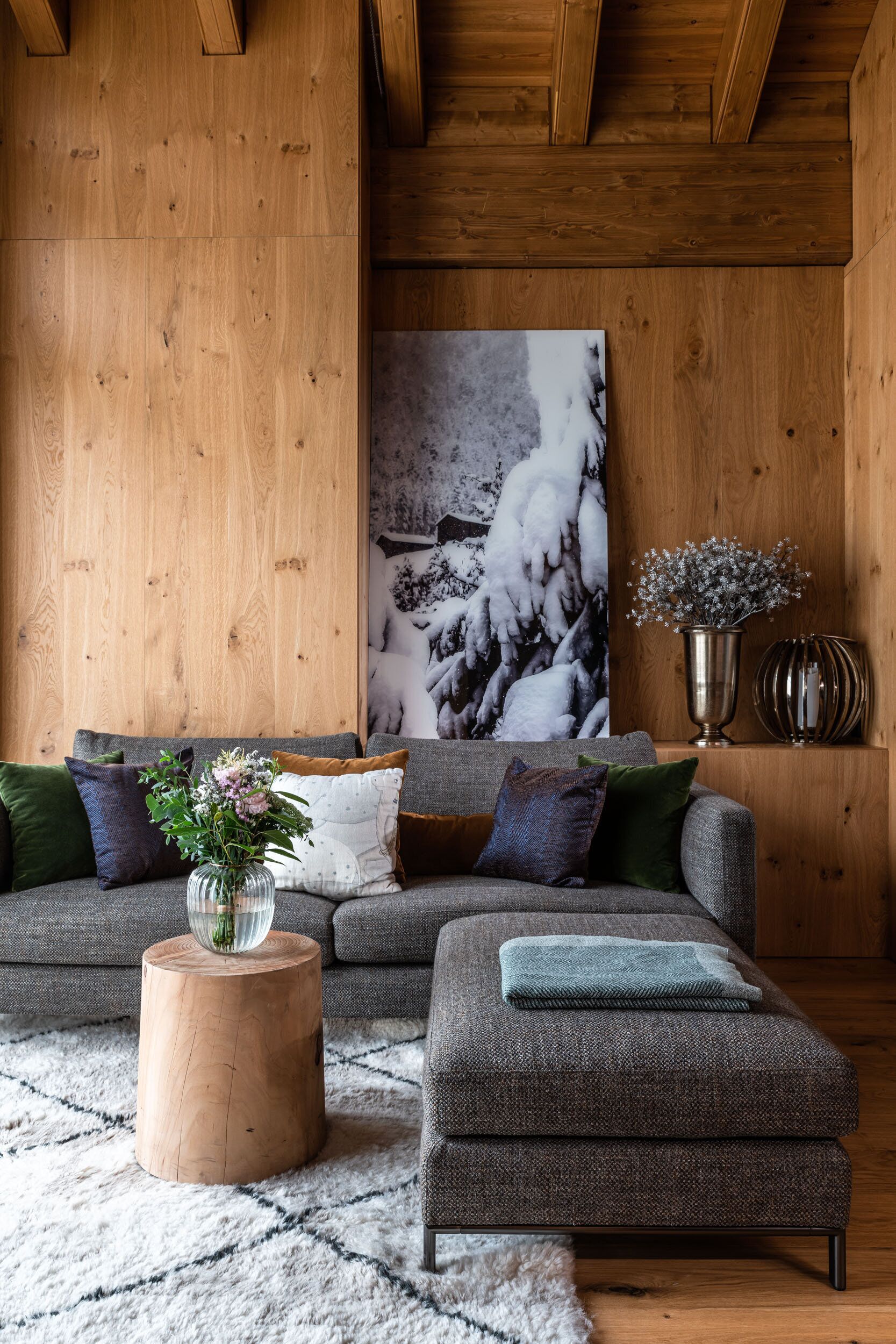 Een gezellige woonkamer met een houten plafond, een groot schilderij van een besneeerd landschap, een grijze bank met veel kussens, een klein houten bijzettafeltje met een bloevas, en een grijze poef.
