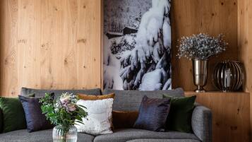 Een gezellige woonkamer met een houten plafond, een groot schilderij van een besneeerd landschap, een grijze bank met veel kussens, een klein houten bijzettafeltje met een bloevas, en een grijze poef.