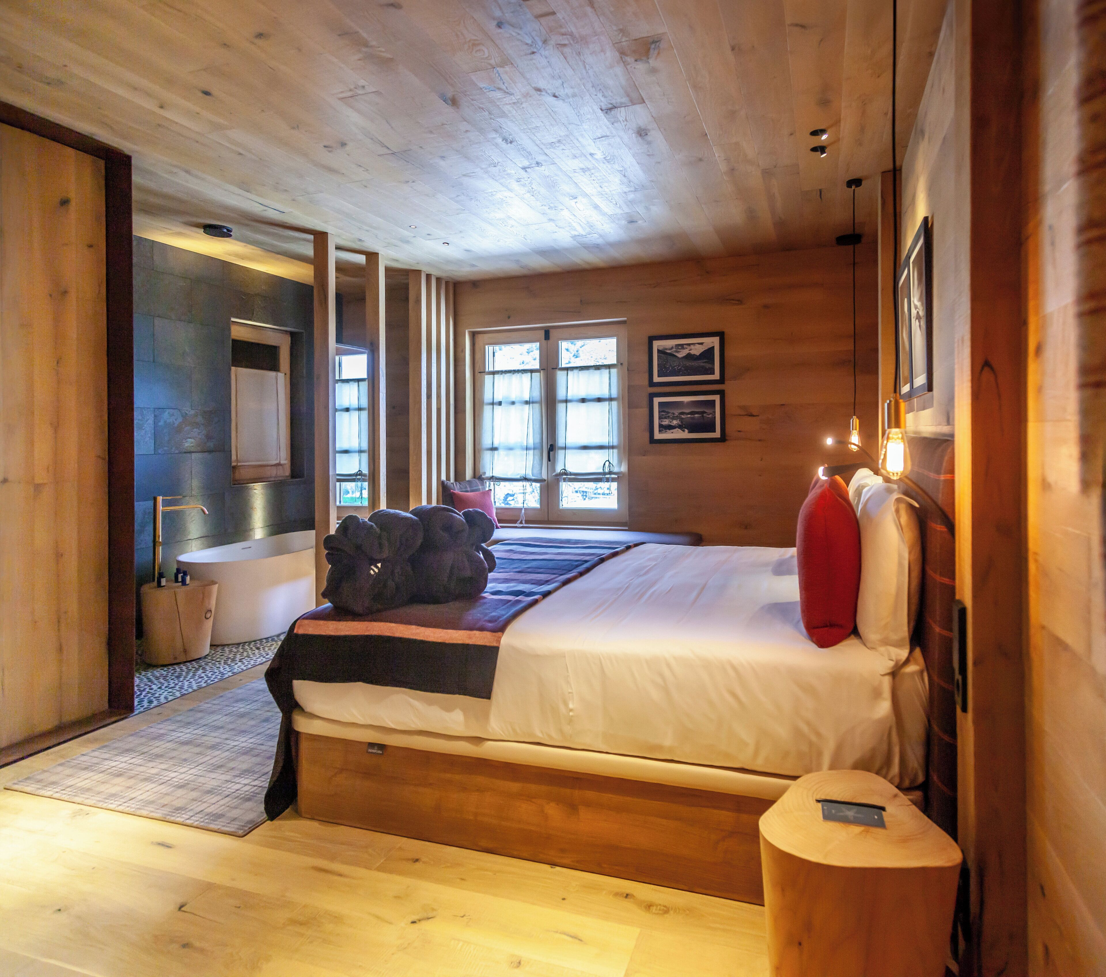 A wooden cabin bedroom with a large bed, a wooden floor, a bathroom with a bathtub, and a window with curtains.