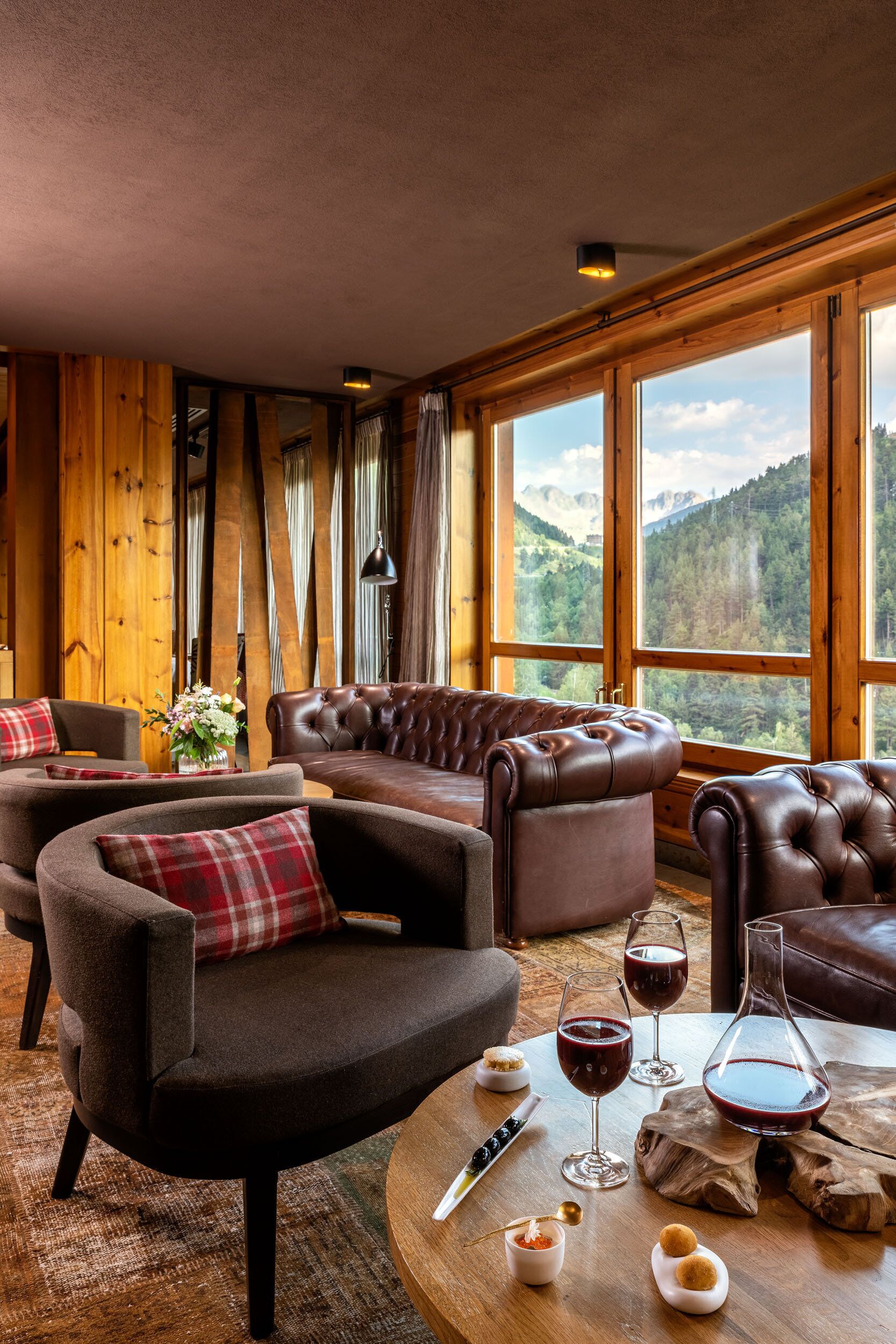 A cozy room with leather sofas, a wooden table with wine glasses, and a view of mountains through large windows.