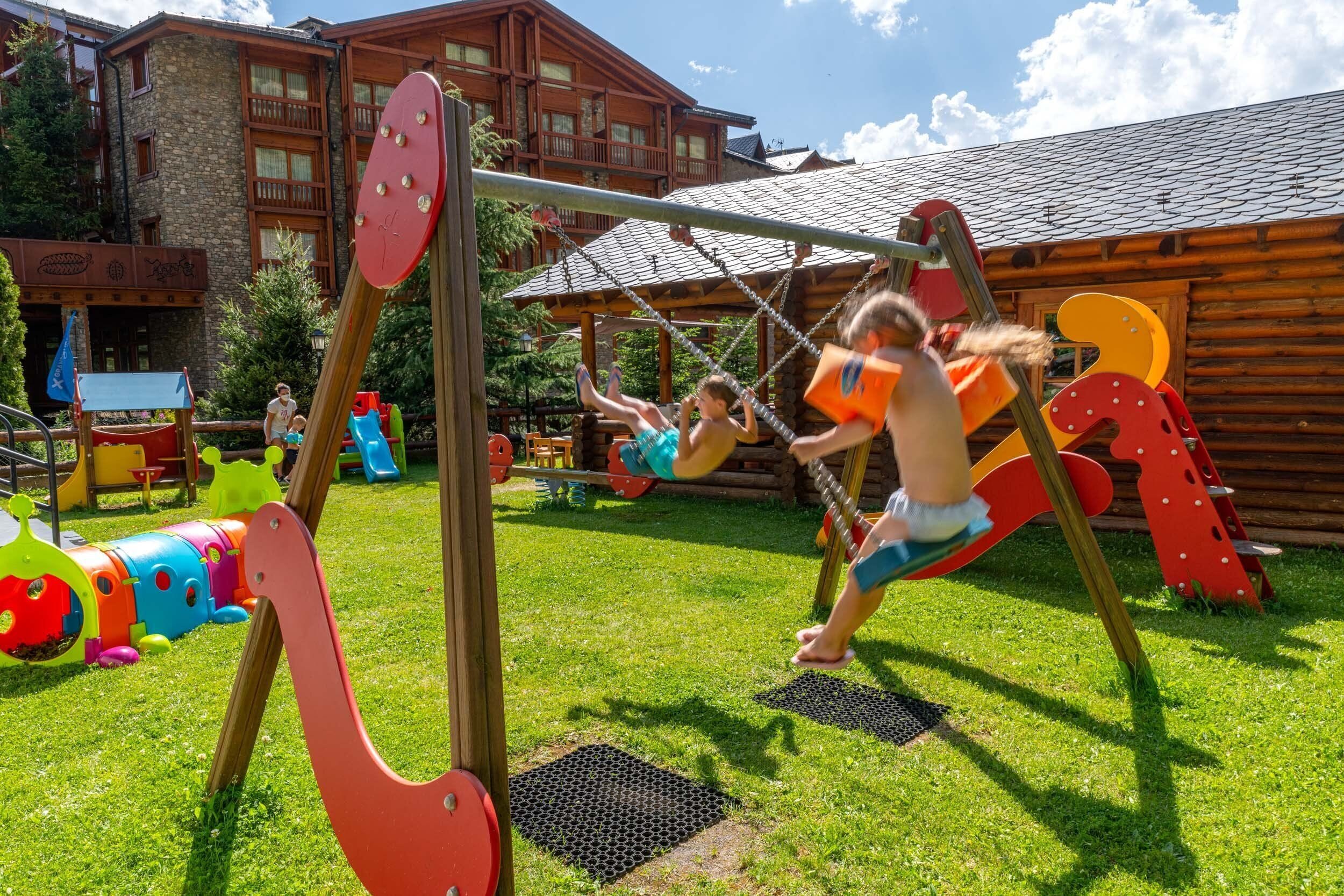 Un parque infantil con columpios y toboganes, rodeado por una cabaña de madera y un edificio con fachada de piedra.