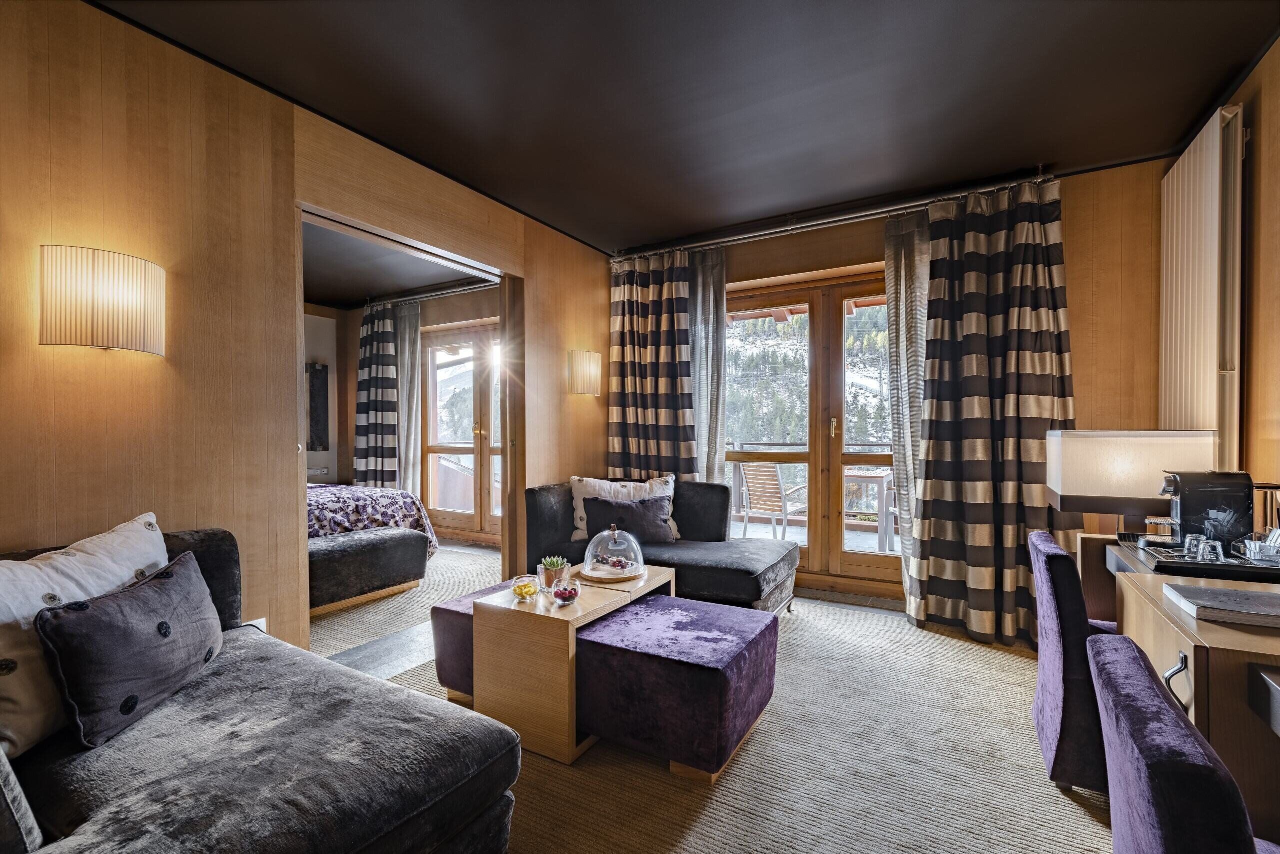 A modern hotel room with a cozy seating area, a wooden desk, and large windows with curtains.