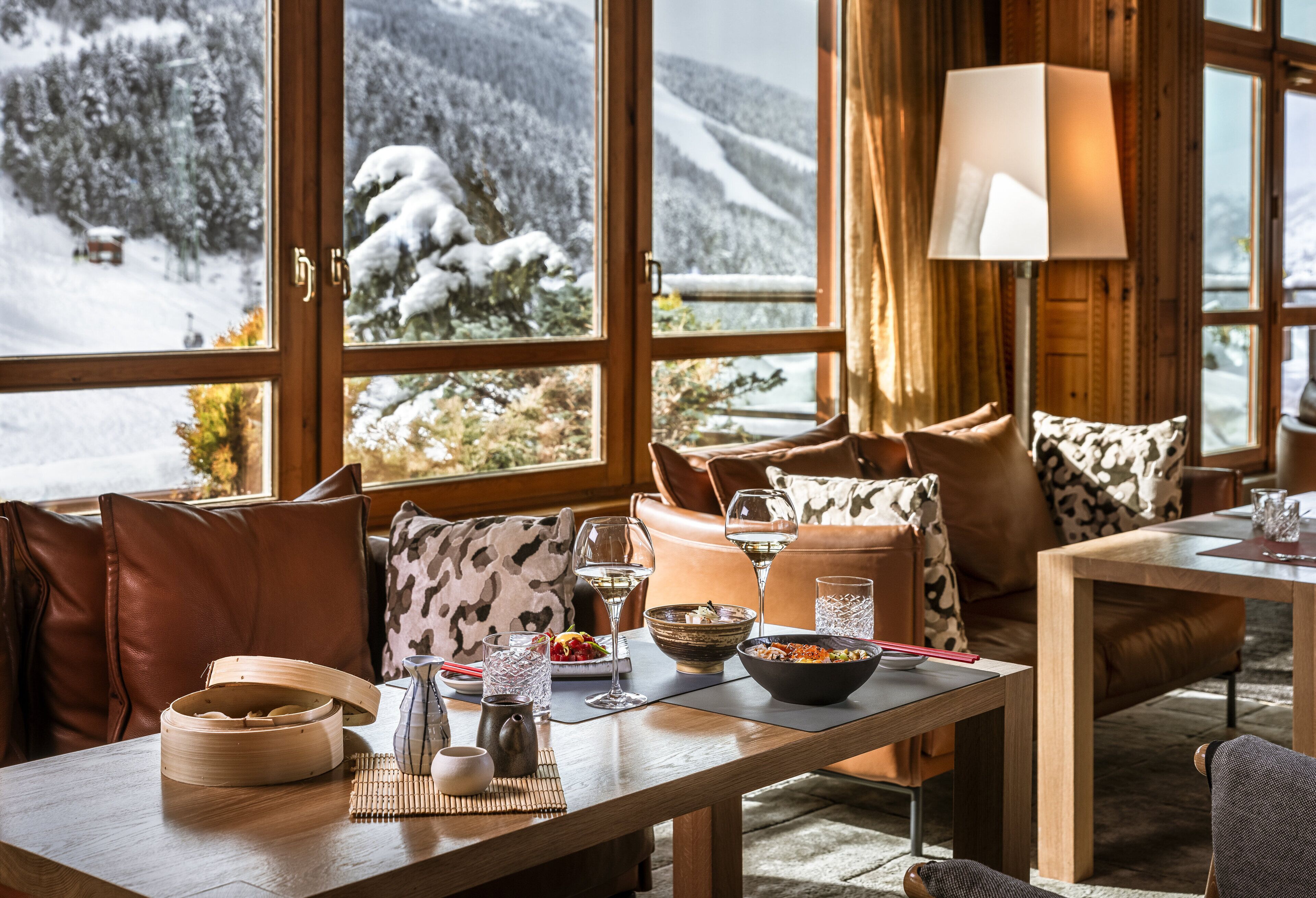 Une pièce chaleureuse avec une grande fenêtre donnant sur un paysage enneigé, une table en bois sur laquelle se trouve un bol de nourriture, et un canapé en cuir agrémenté de coussins à motifs.
