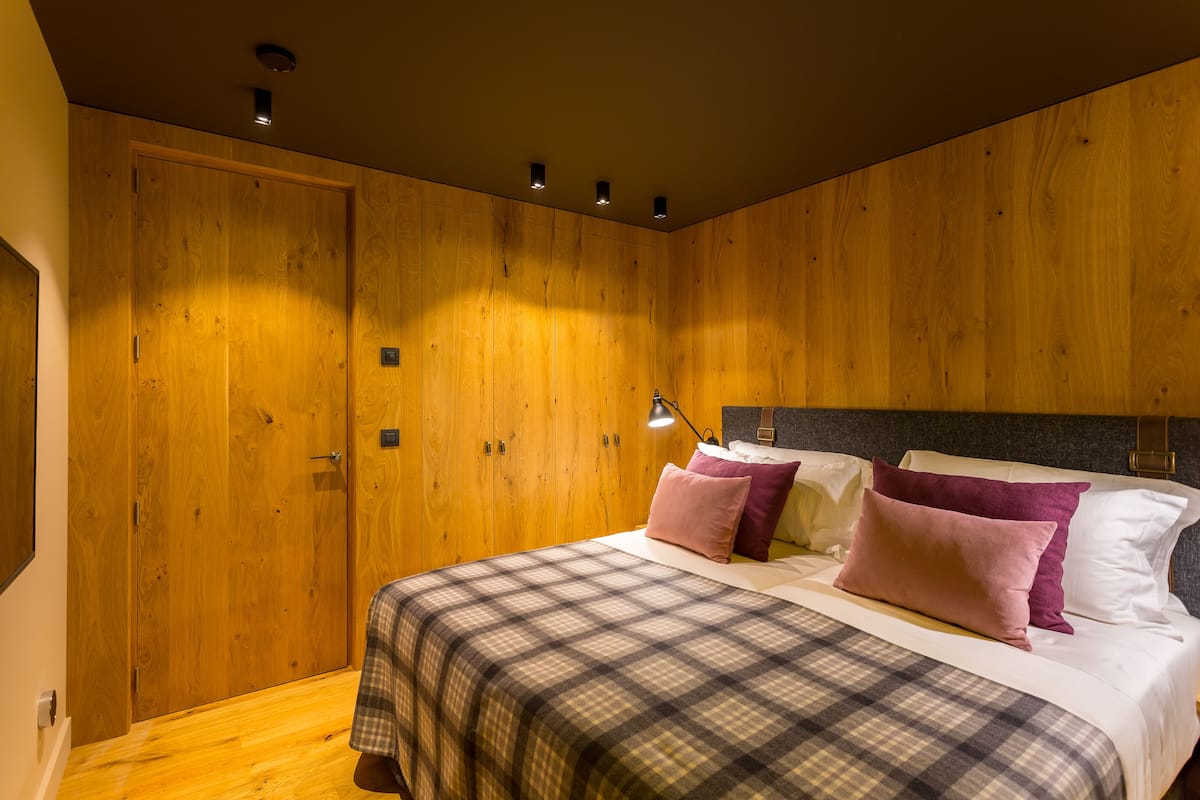 A bedroom with a wooden wall, a bed with a plaid bedspread, and two bedside lamps.