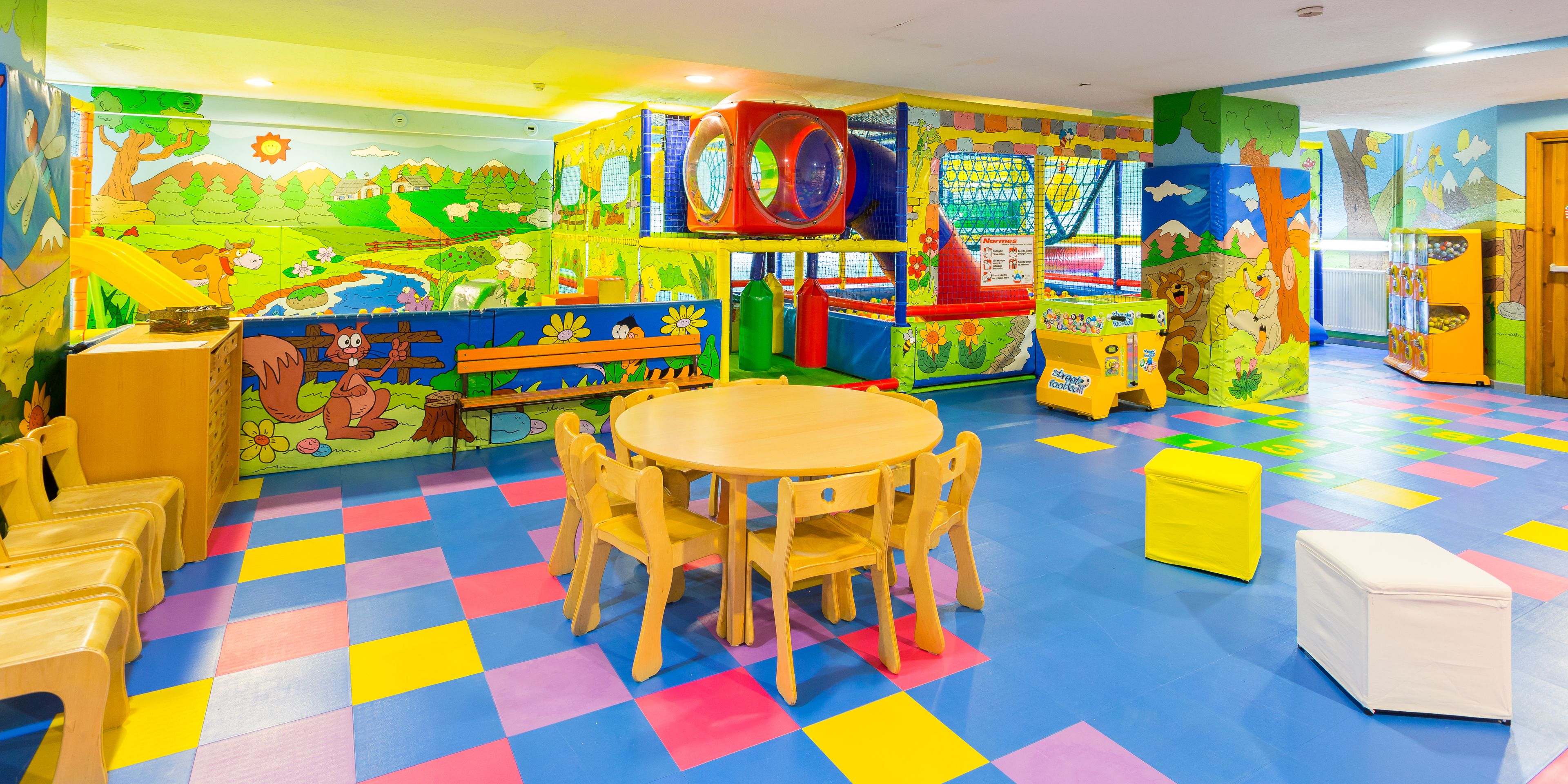 A children's play area with colorful flooring, play structures, and a round wooden table with chairs.