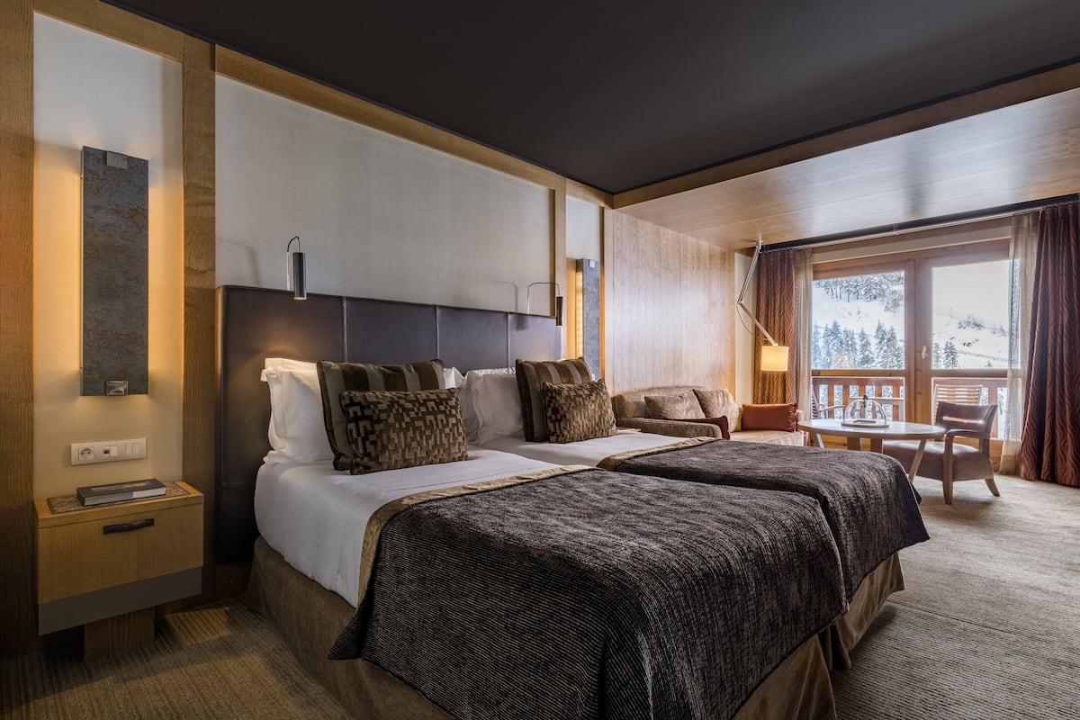 A hotel room with two beds, a wooden headboard, a small table, and a balcony with a view of a snowy landscape.