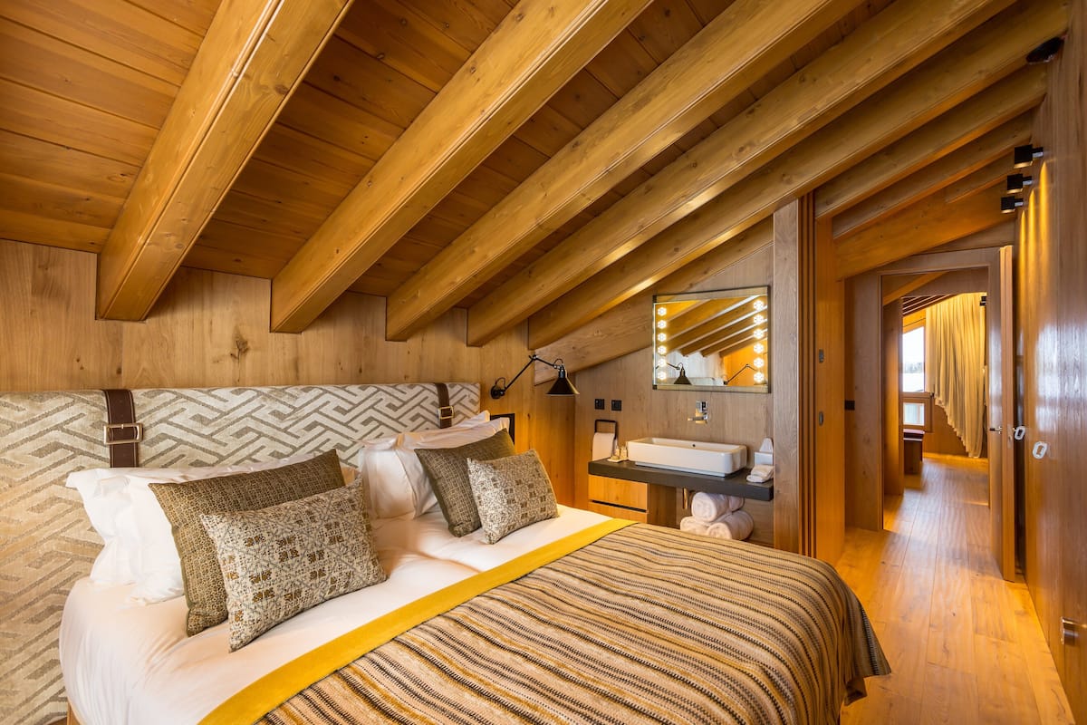 A wooden ceiling with exposed beams, a bed with patterned bedding, a bedside table, a mirror with lights, and a bathroom with a sink and towel rack.