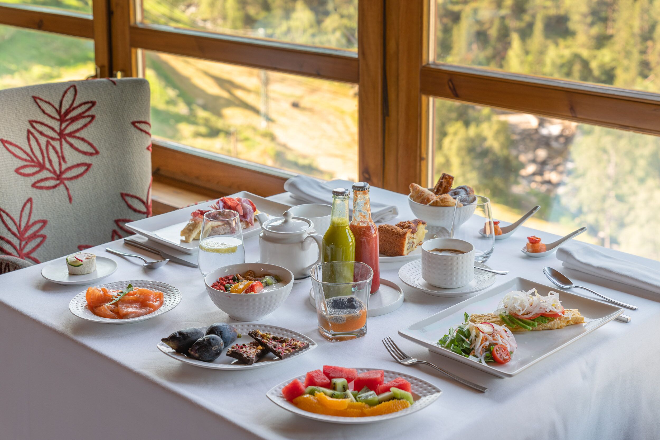 Une table de petit-déjeuner dressée avec divers plats, notamment du saumon fumé, des fruits et des viennoiseries, le tout mis en valeur par un paysage pittoresque en arrière-plan.