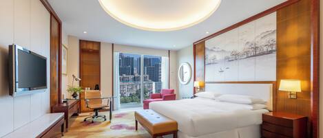 Ein Hotelzimmer mit einem großen Bett, einem Schreibtisch, einem Sessel, einem Fernseher und Blick auf die Stadt.