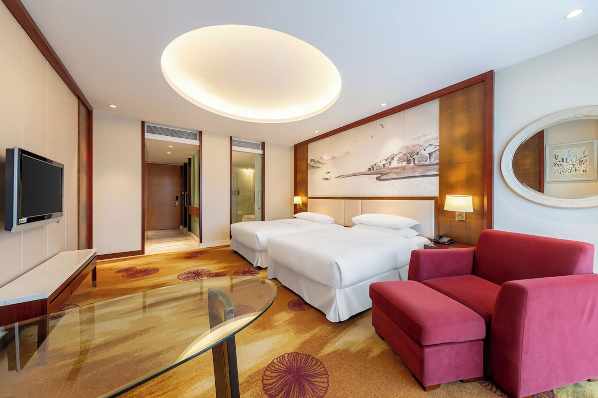 Habitación de hotel con dos camas, un televisor, una mesa de vidrio, un sofá rojo y un cuadro grande en la pared.