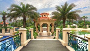 Eine Brücke führt zu einem Gazebo mit einem Ziegeldach, umgeben von Palmen und einem Pool.