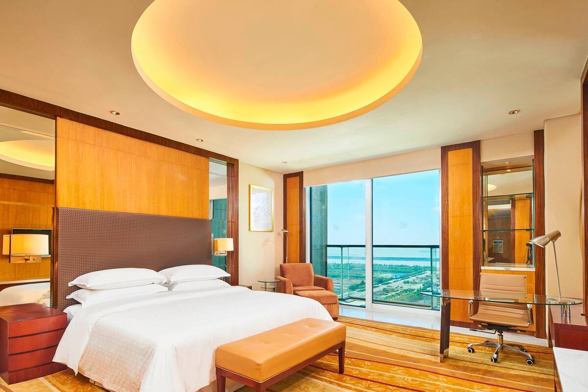 Habitación de hotel con una cama grande, un escritorio con una silla, un banco y un ventanal con vistas a la ciudad.