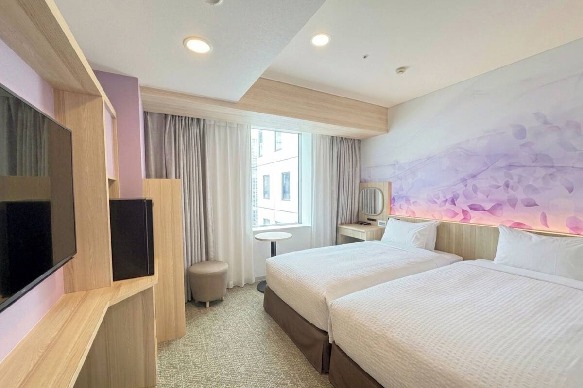 Habitación de hotel con dos camas, televisión, una mesita, una silla y ventana con cortinas.