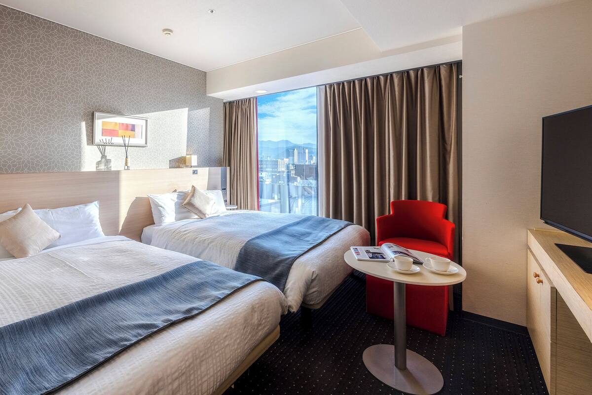 Una habitación de hotel con dos camas, una silla roja, una mesita con tazas y un televisor.