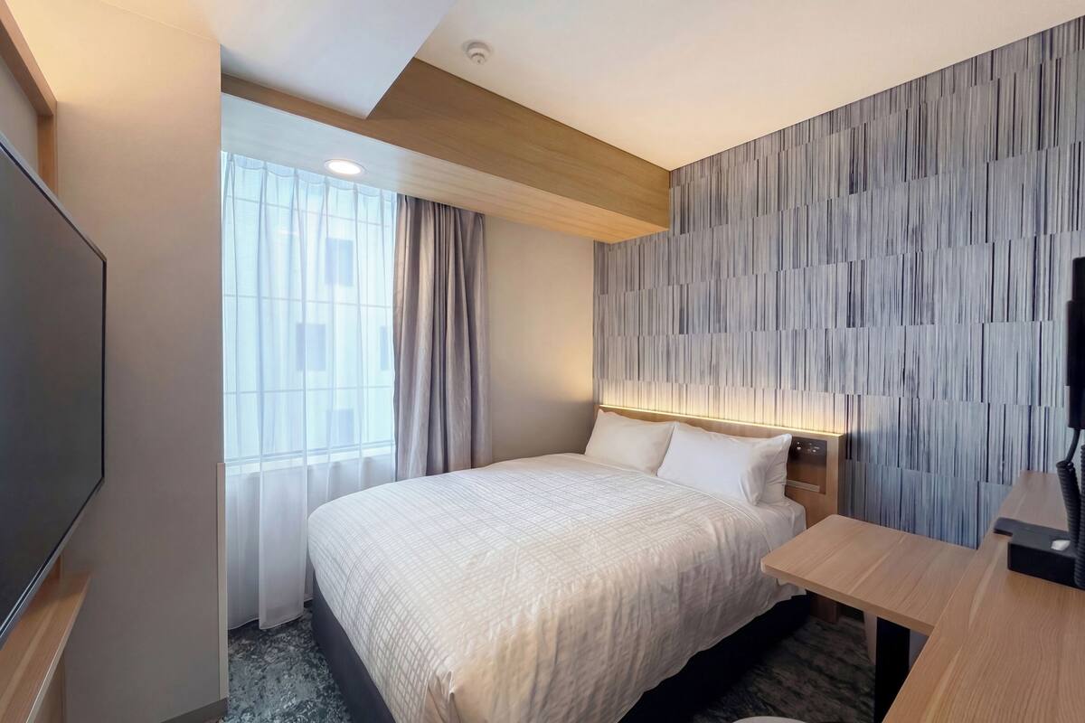 Una habitación de hotel con cama, escritorio, televisor y ventana con cortinas.