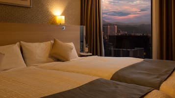A hotel room with two beds, a nightstand with a lamp, and a view of a cityscape at dusk.