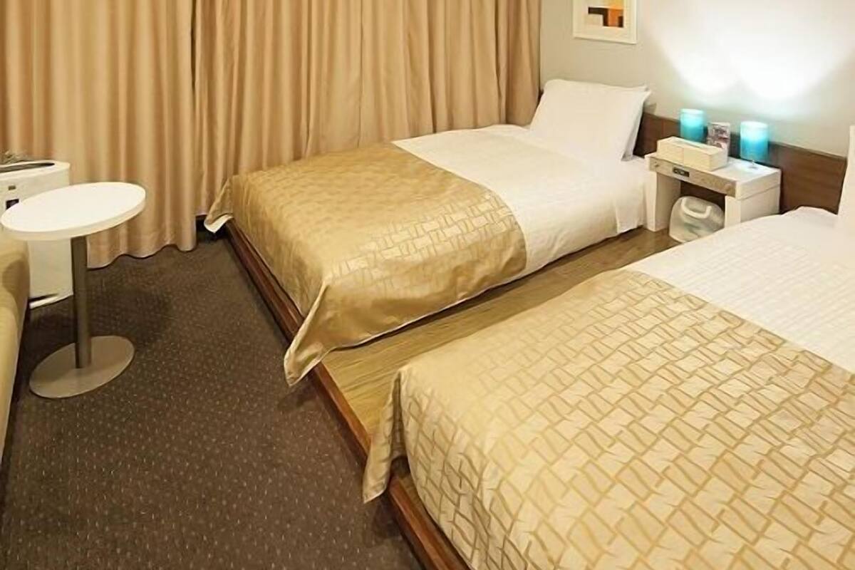 Habitación de hotel con dos camas, una mesita redonda pequeña, una mesita de noche y una lámpara de pared.