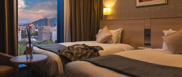 Habitación de hotel con dos camas, vista a la ciudad, una lámpara y una botella de perfume sobre una mesita.