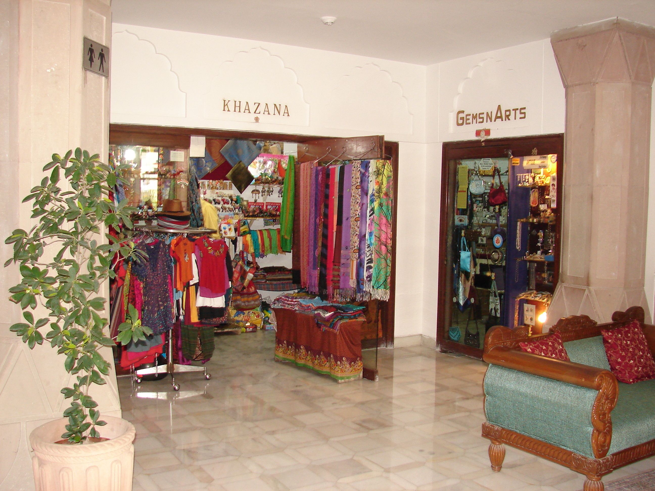 Un elegante vestíbulo de hotel boutique con una zona de estar, una planta y dos tiendas llamadas 'KHAZANA' y 'GEMS N ARTS'.