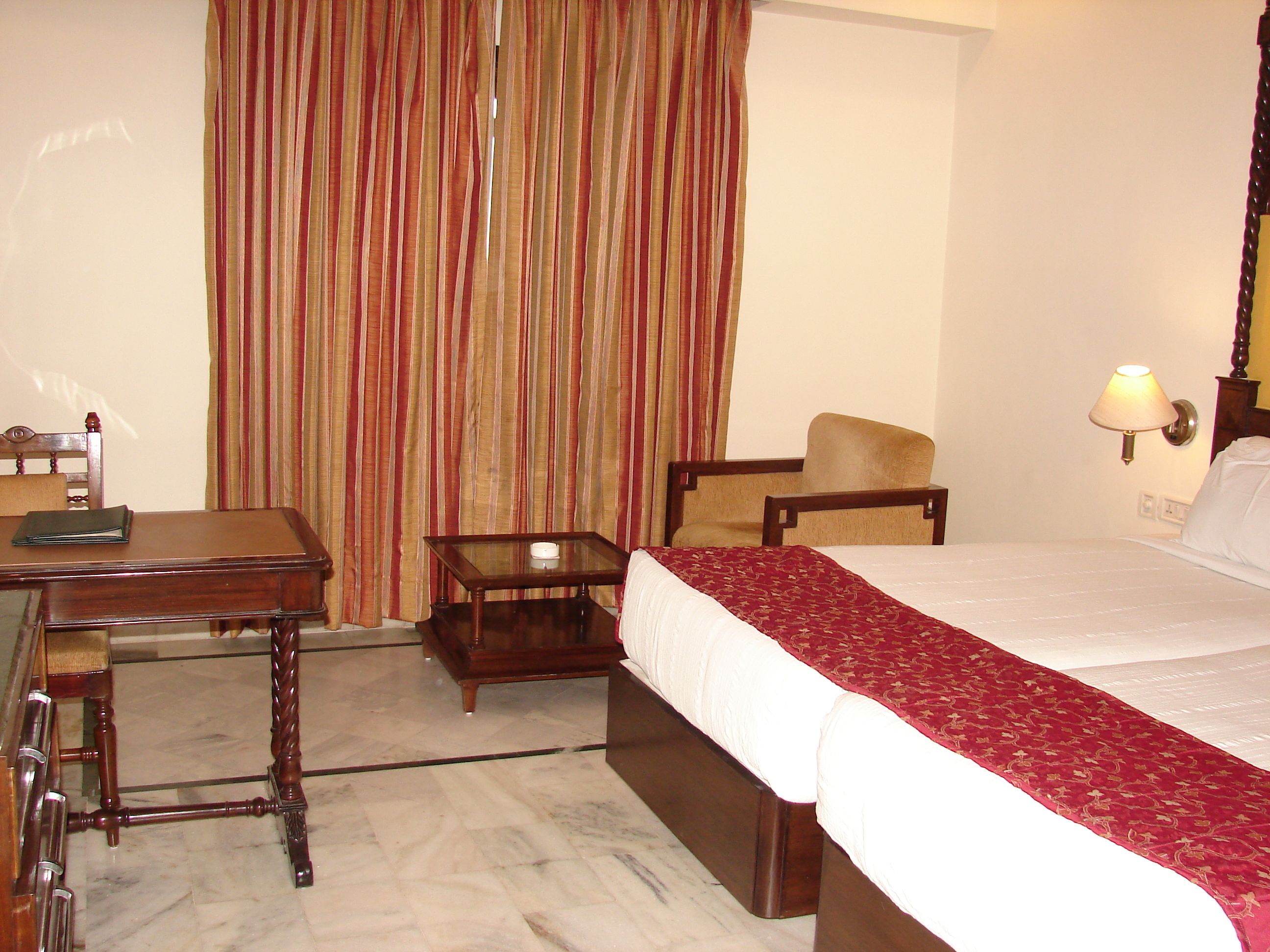 A hotel room with a bed, a desk, a chair, a small table, and a lamp.