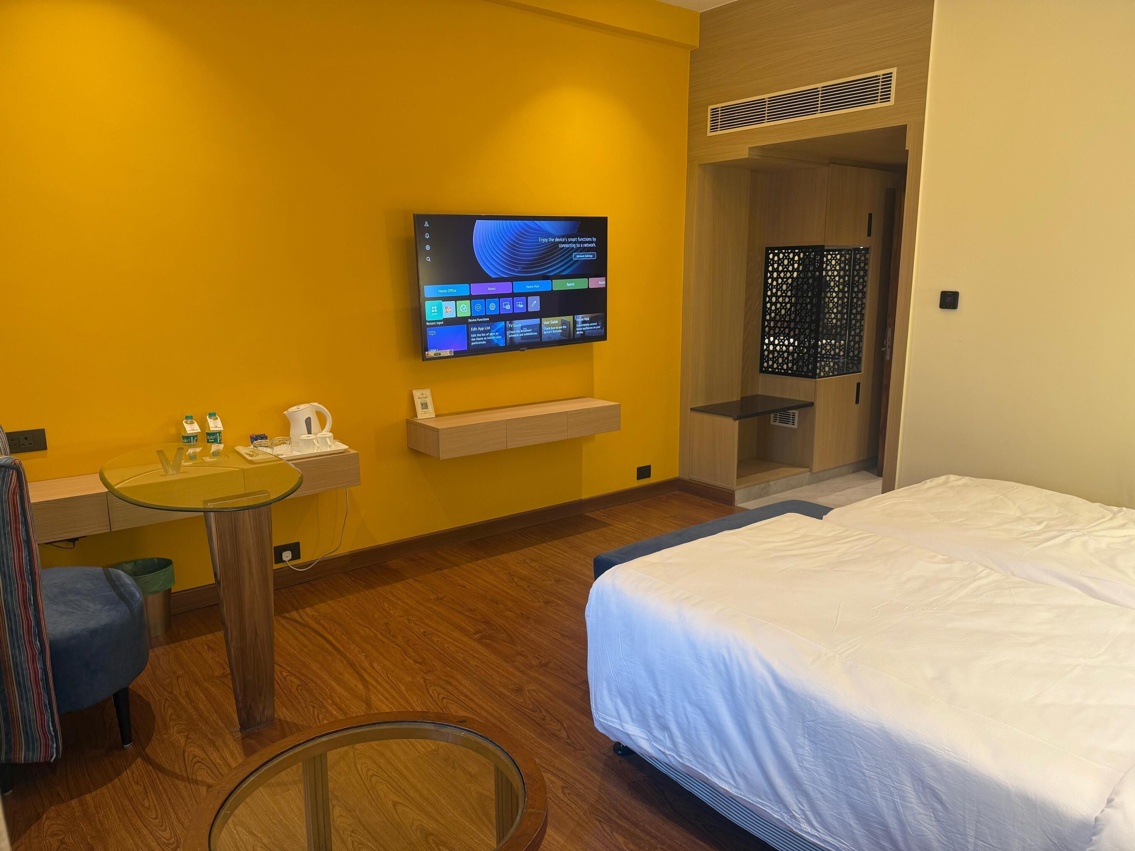 A hotel room with a bed, a desk with a chair, a television mounted on the wall, and a small shelf with a kettle and cups.