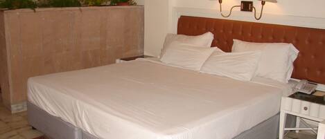 A neatly made bed with white linens, a brown headboard, and a bedside table with a lamp and a telephone.