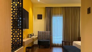Habitación de hotel con piso de madera, un escritorio con silla, cama, televisor montado en la pared y ventana con cortinas.