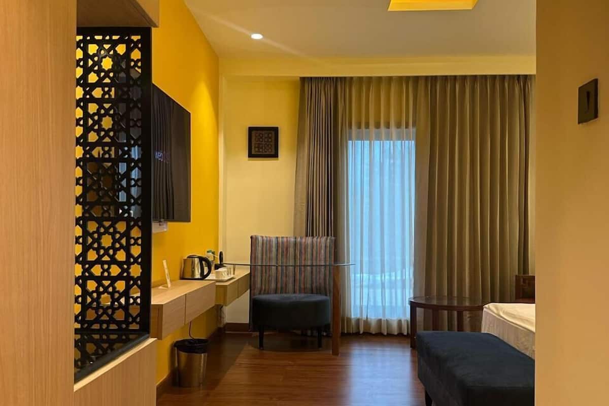 Habitación de hotel con piso de madera, un escritorio con silla, cama, televisor montado en la pared y ventana con cortinas.
