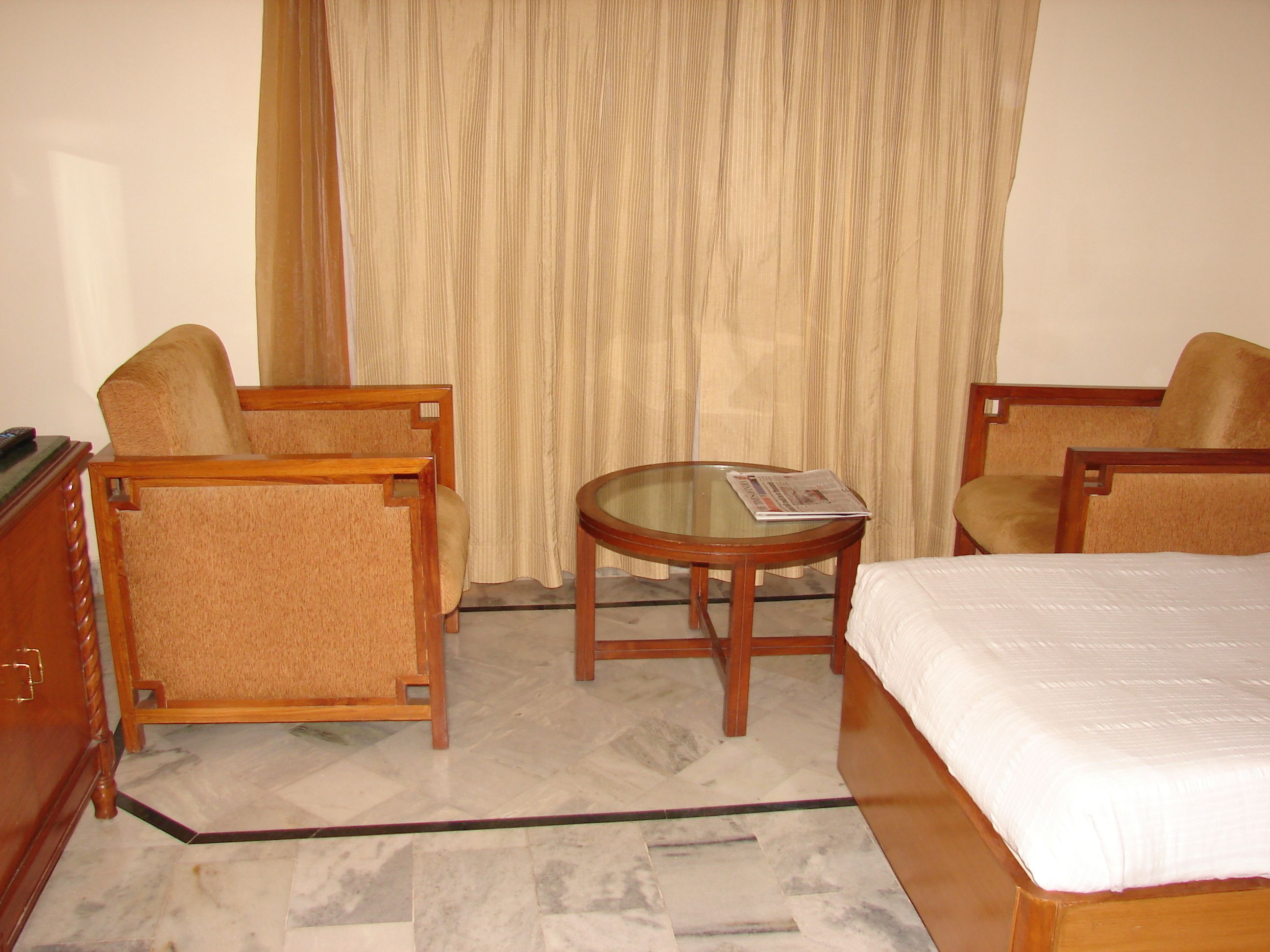 Una habitación de hotel con una cama, dos sillones, una mesita redonda con un periódico y un aparador de madera.