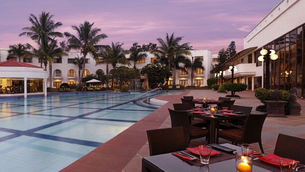 A poolside dining area with tables and chairs, a pool, and a building with multiple balconies and palm trees. - Lemon Tree Hotel, Aurangabad (Chhatrapati Sambhajinagar)