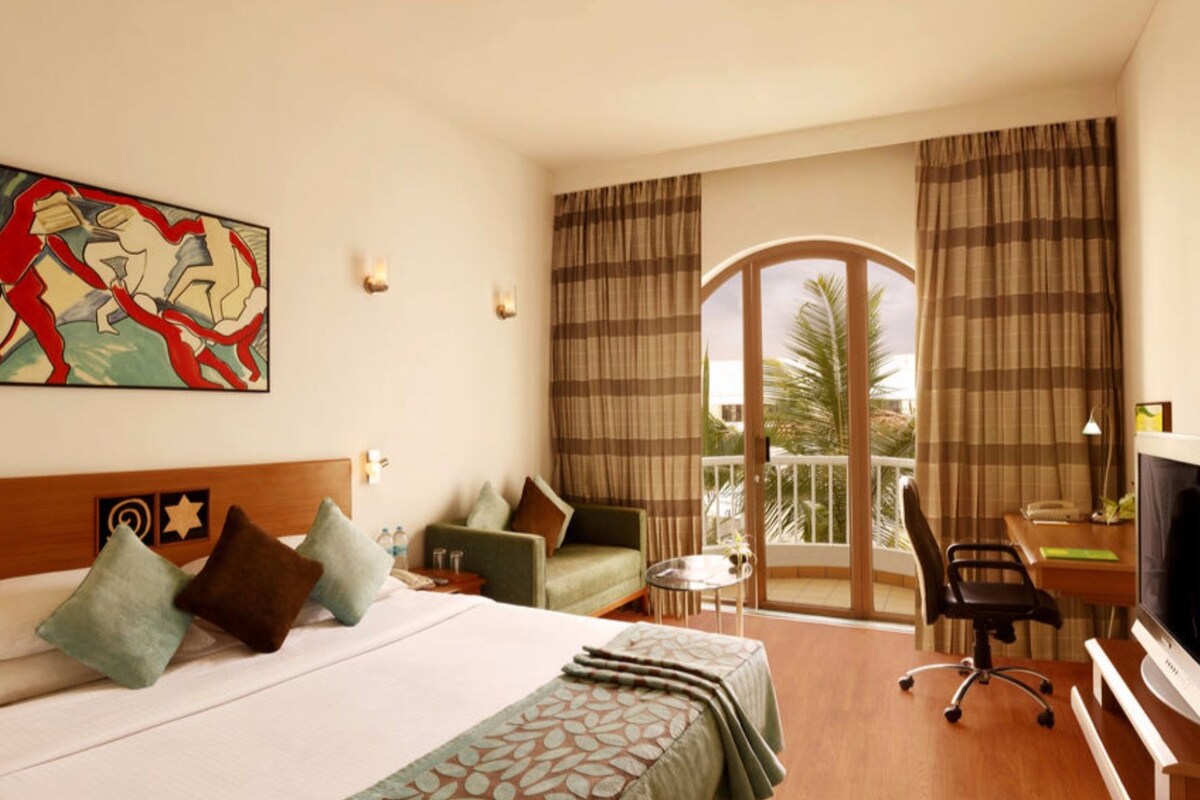 A hotel room with a bed, a desk with a computer, a chair, a TV, and a balcony with curtains.