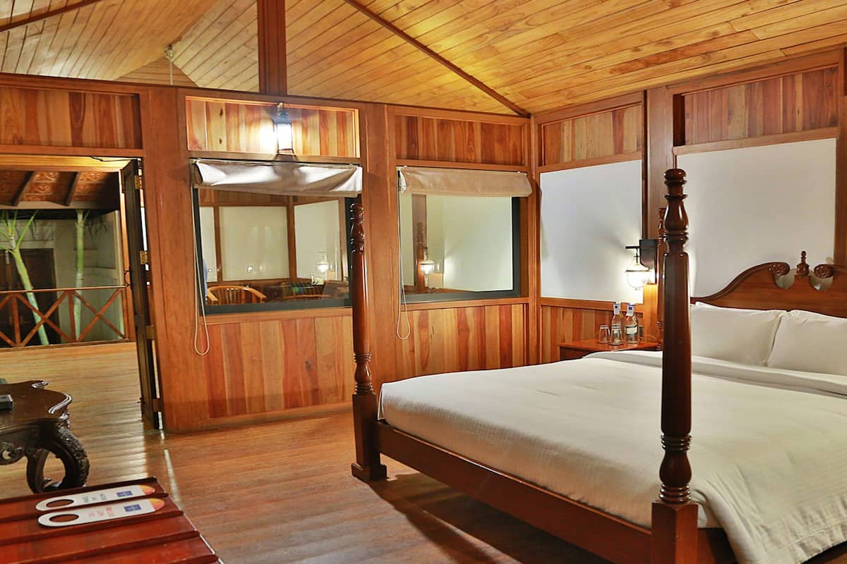 Uma cama de madeira com dossel, roupa de cama branca, uma mesa de cabeceira de madeira com abajur e janela de madeira com vista para o exterior.