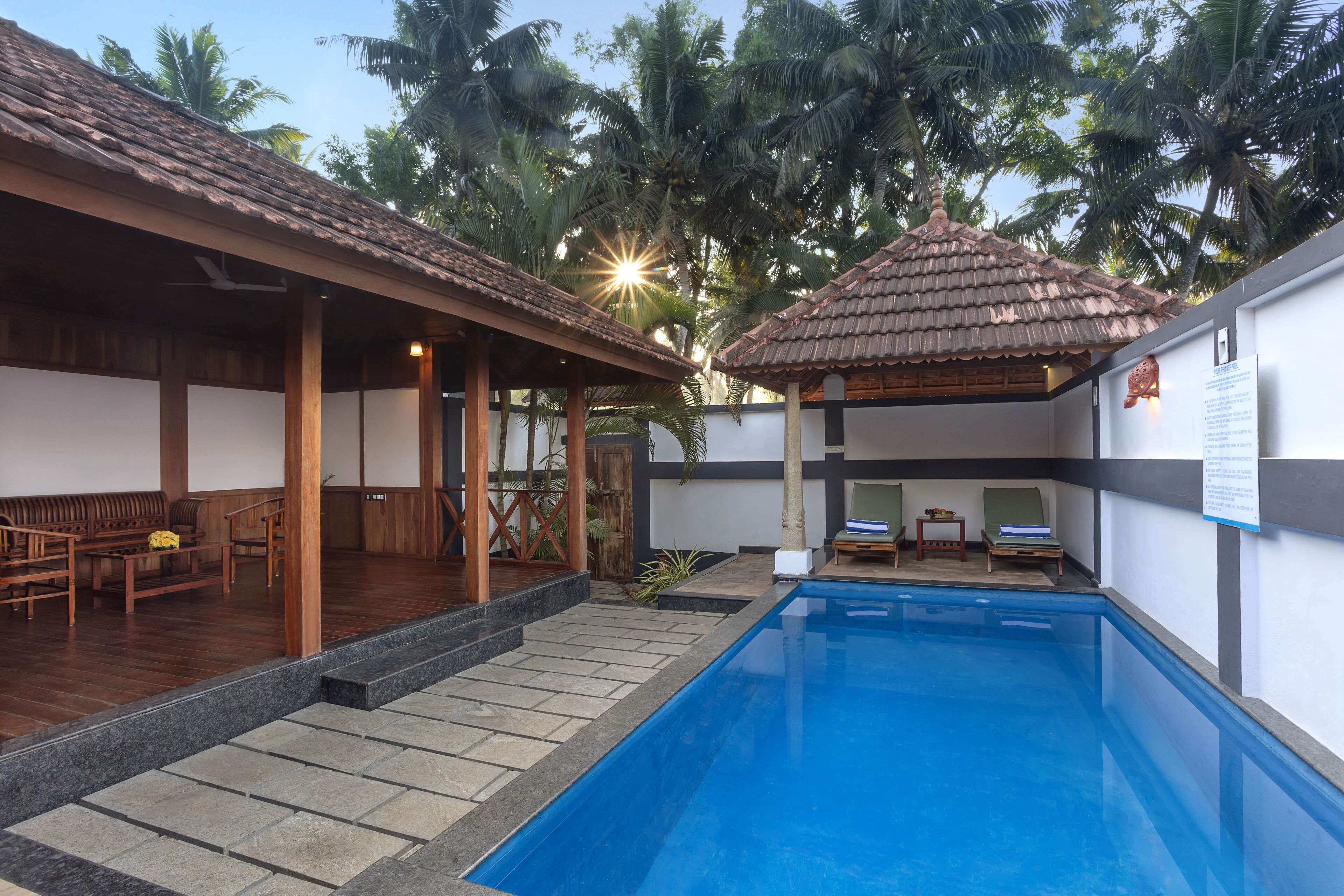 Zona de piscina con solárium de baldosas, asientos de lounge de madera y un área de estar cubierta con techo de baldosas.