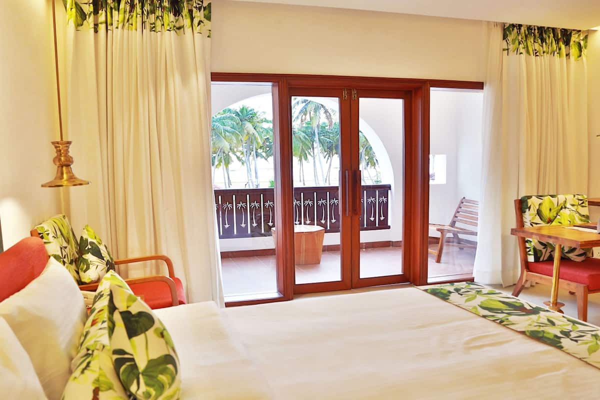 Quarto de hotel com cama, escrivaninha, cadeira e varanda com vista para palmeiras.