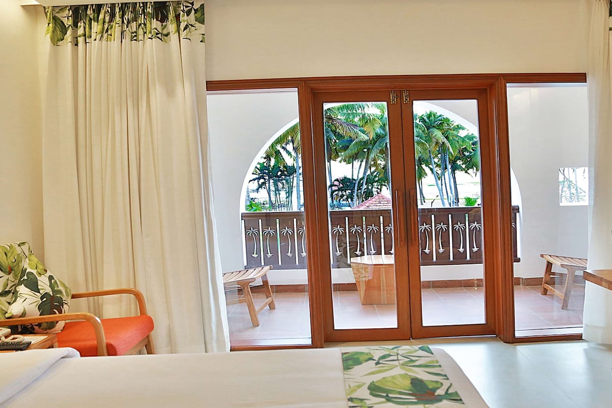 Habitación de hotel con cama, escritorio, una silla y balcón con vistas a palmeras.