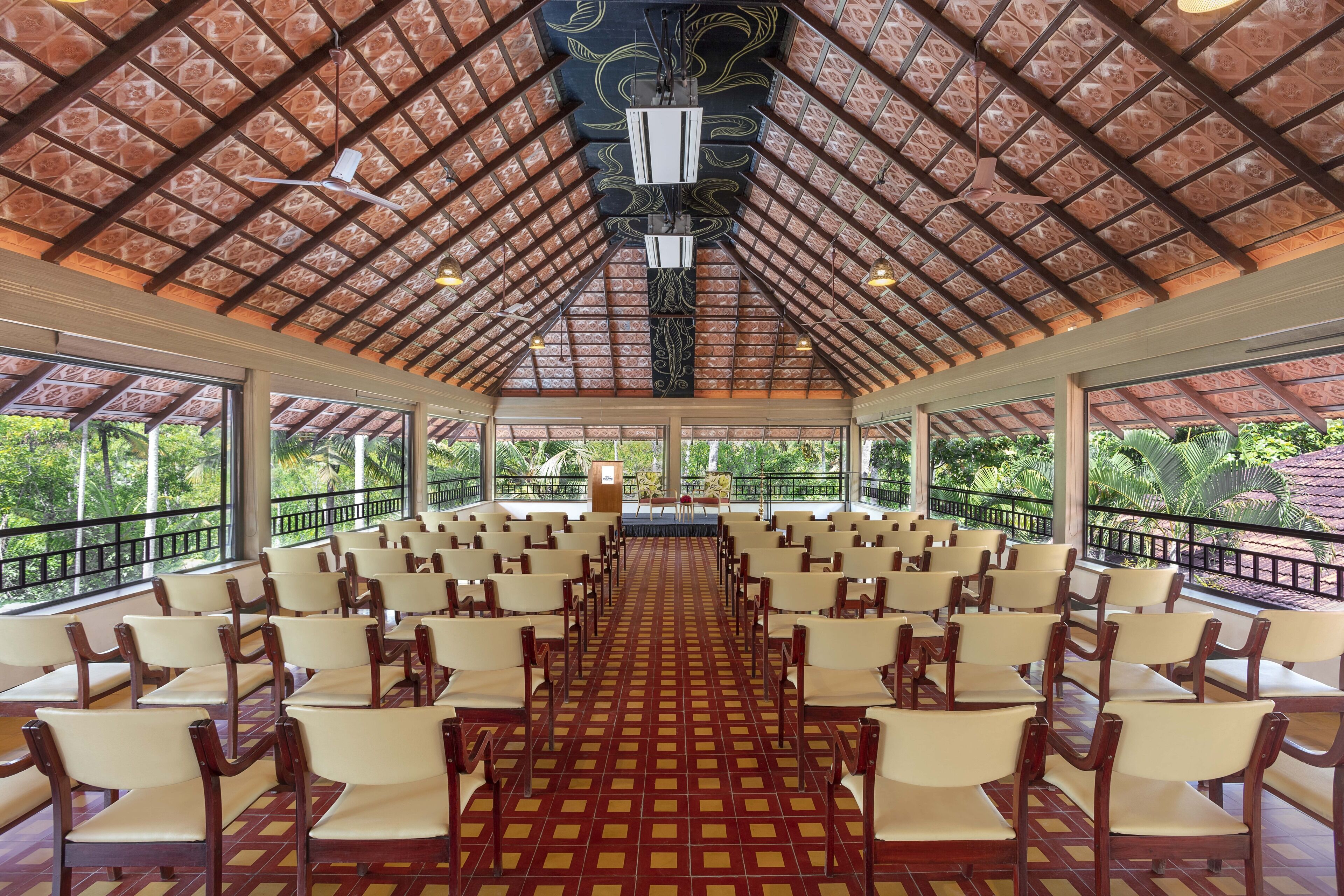 Un salón amplio con filas de sillas y un escenario, ventanas grandes y un techo con diseño.