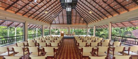 Un salón amplio con filas de sillas y un escenario, ventanas grandes y un techo con diseño.