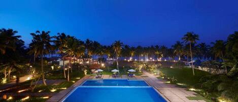 Vista nocturna de una piscina rectangular con plataforma empedrada, rodeada de palmeras e iluminada con luz ambiental.