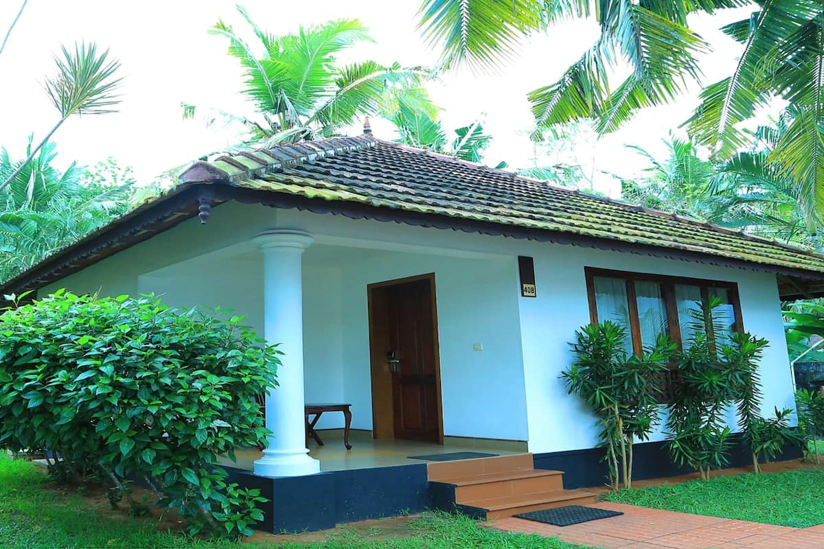 Uma pequena casa térrea com telhado de telhas, varanda com um banco e um pequeno jardim com palmeiras e arbustos.
