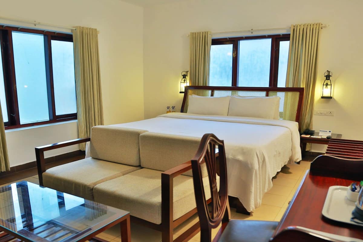 Quarto de hotel com uma cama grande, duas cadeiras, uma mesa de vidro e duas janelas com cortinas.