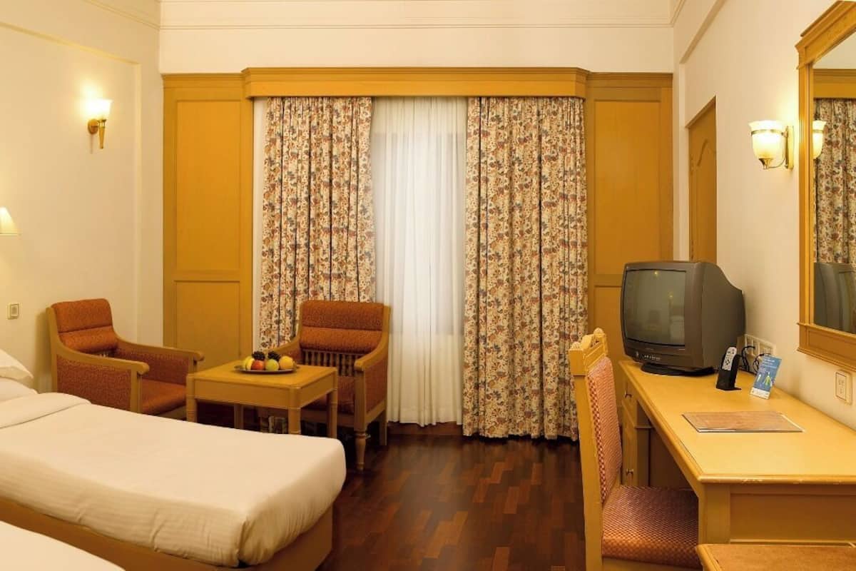Quarto de hotel com duas camas de solteiro, uma escrivaninha com televisão, uma cadeira, uma pequena mesa com frutas e uma janela com cortinas florais.