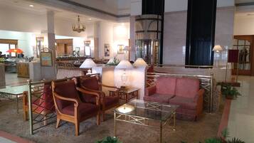 A spacious lobby with a seating area featuring a glass coffee table, two wooden armchairs, and a patterned sofa. A chandelier hangs from the ceiling, and there is a large mirror on the wall.
