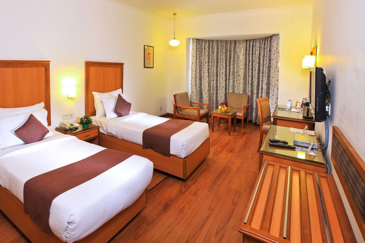 Quarto de hotel com duas camas, uma escrivaninha, uma cadeira, televisão e janela com cortinas.