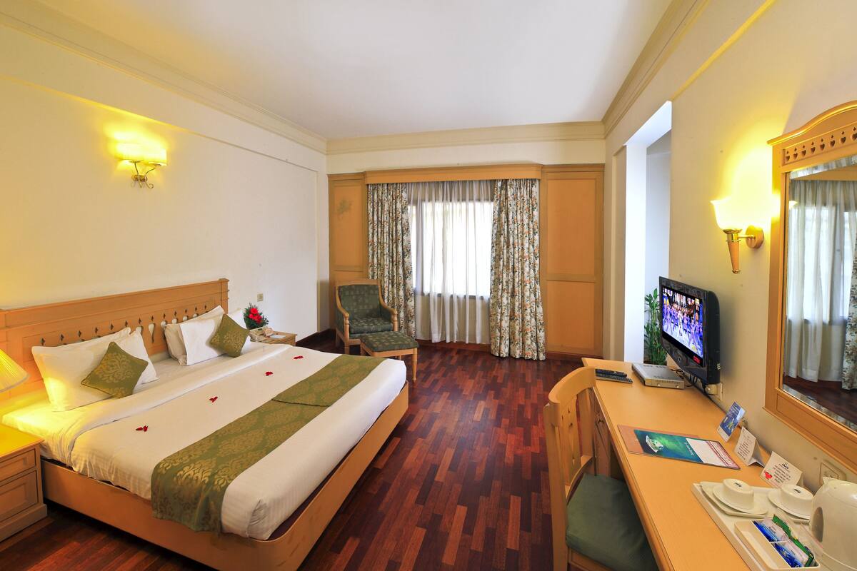 Quarto de hotel com cama, mesa de cabeceira, cadeira, escrivaninha, televisão e uma janela grande com cortinas.