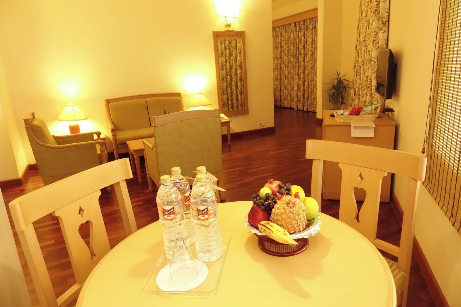 Habitación de hotel con una mesa de comedor dispuesta con botellas de agua, un frutero y un plato. Hay sillas alrededor de la mesa, un televisor en la pared y una ventana con cortinas.