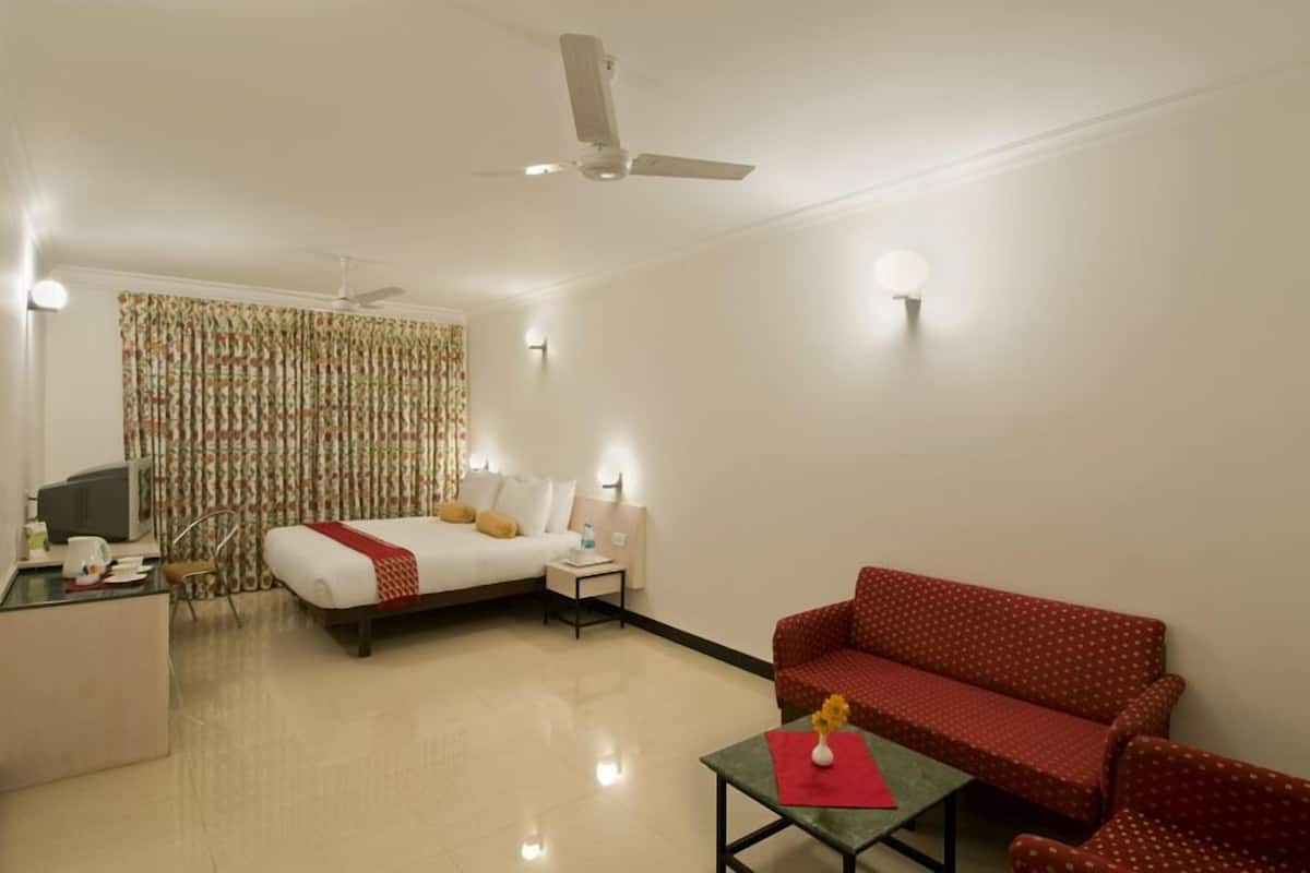 A hotel room with a bed, a red sofa, a small table with a vase, a desk with a computer, and a ceiling fan.