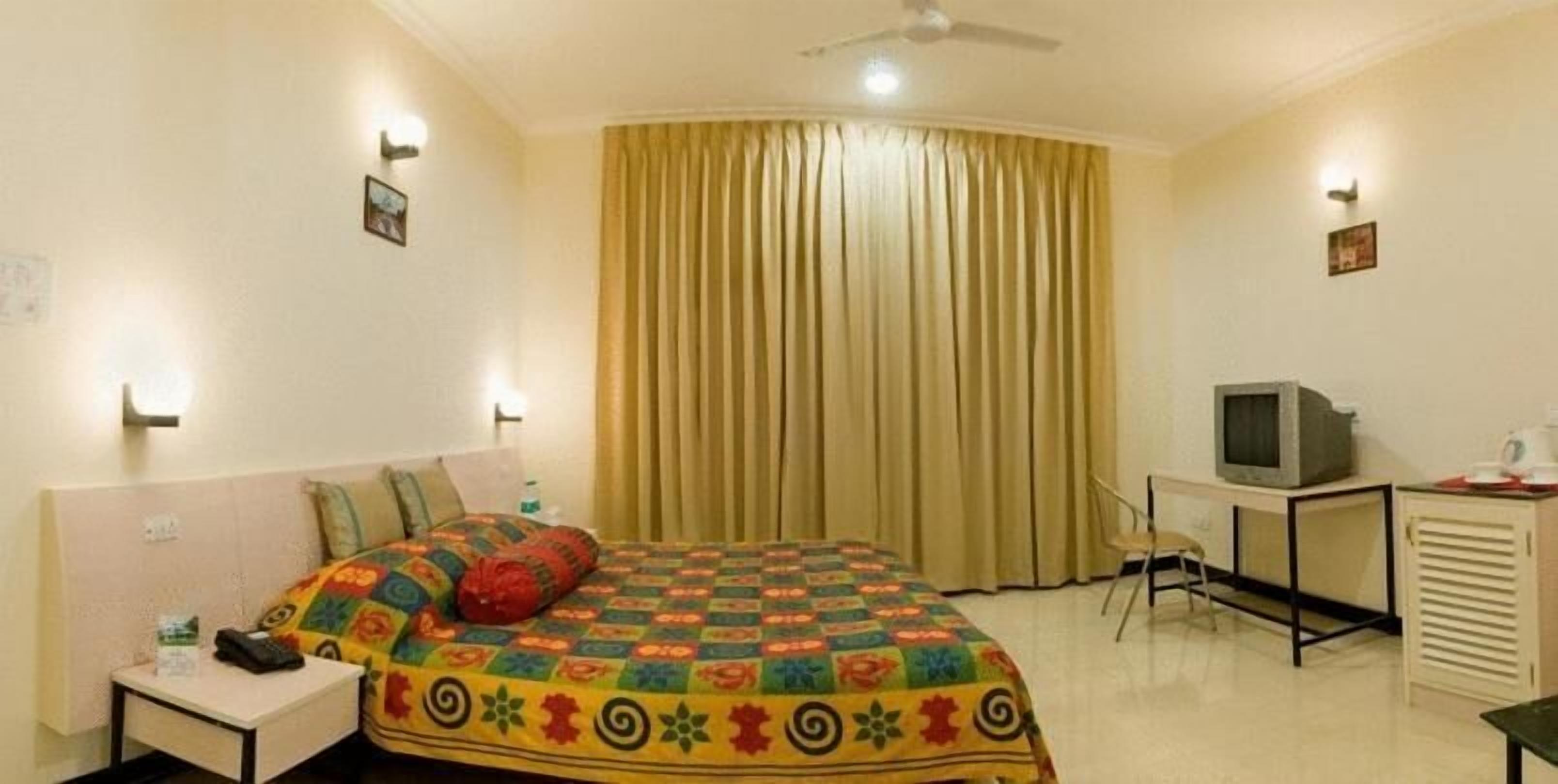 Quarto de hotel com cama, escrivaninha, cadeira, televisão e cortinas.
