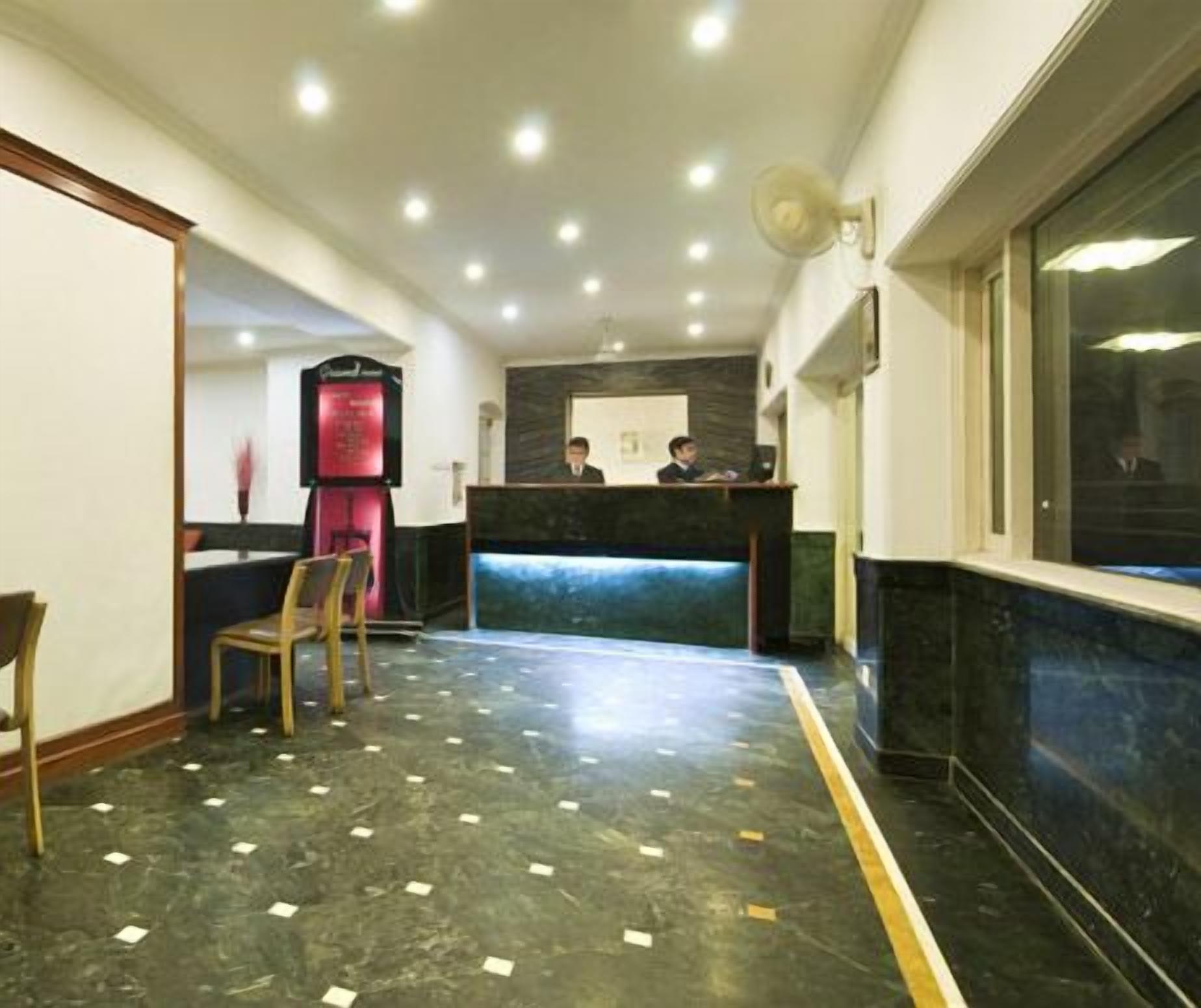 A hotel reception area with a reception desk, two staff members, a seating area with chairs, and a marble floor.